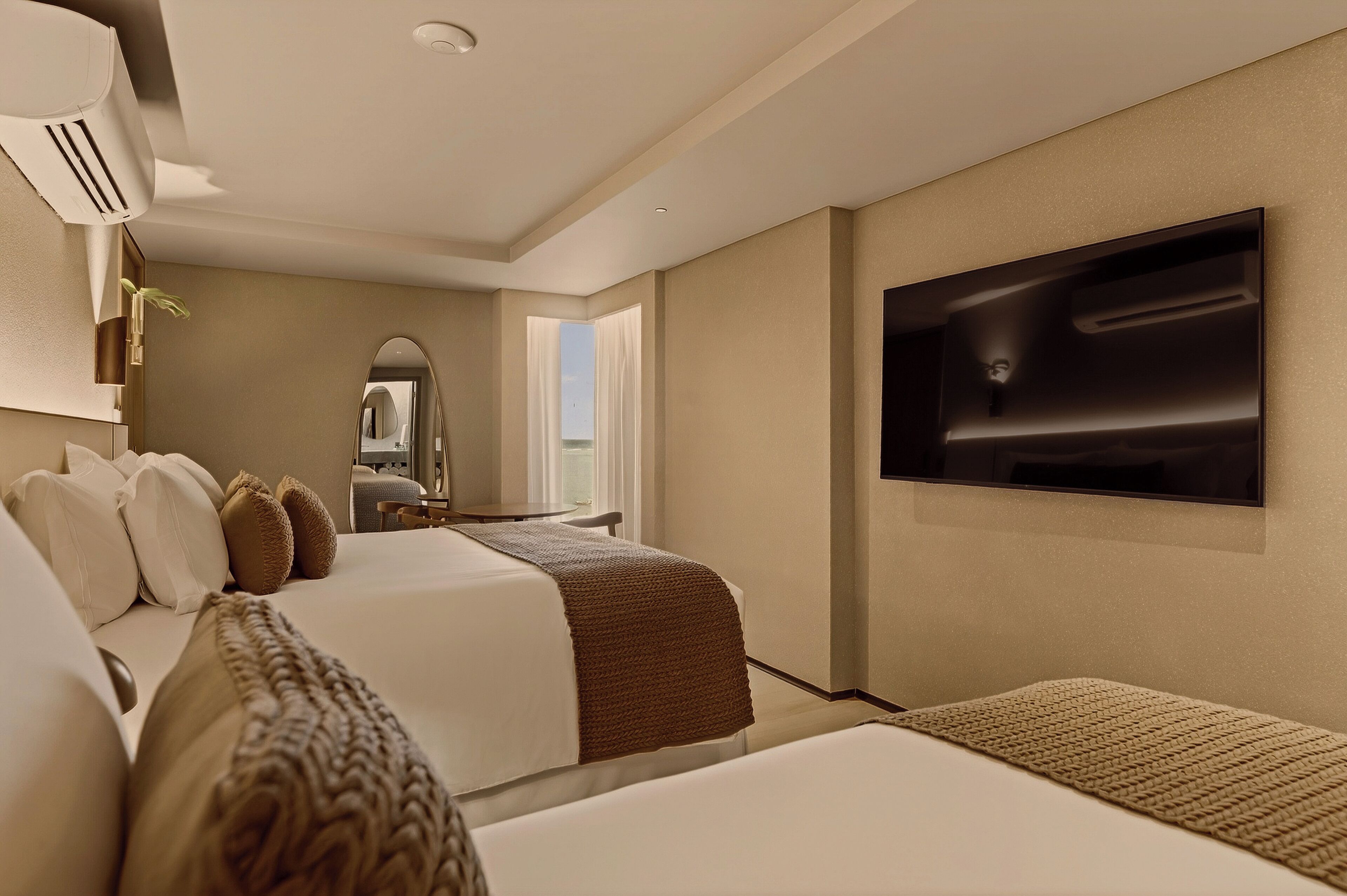 Habitación de hotel con dos camas, un televisor de pantalla plana montado en la pared, un espejo grande y una ventana con cortinas.