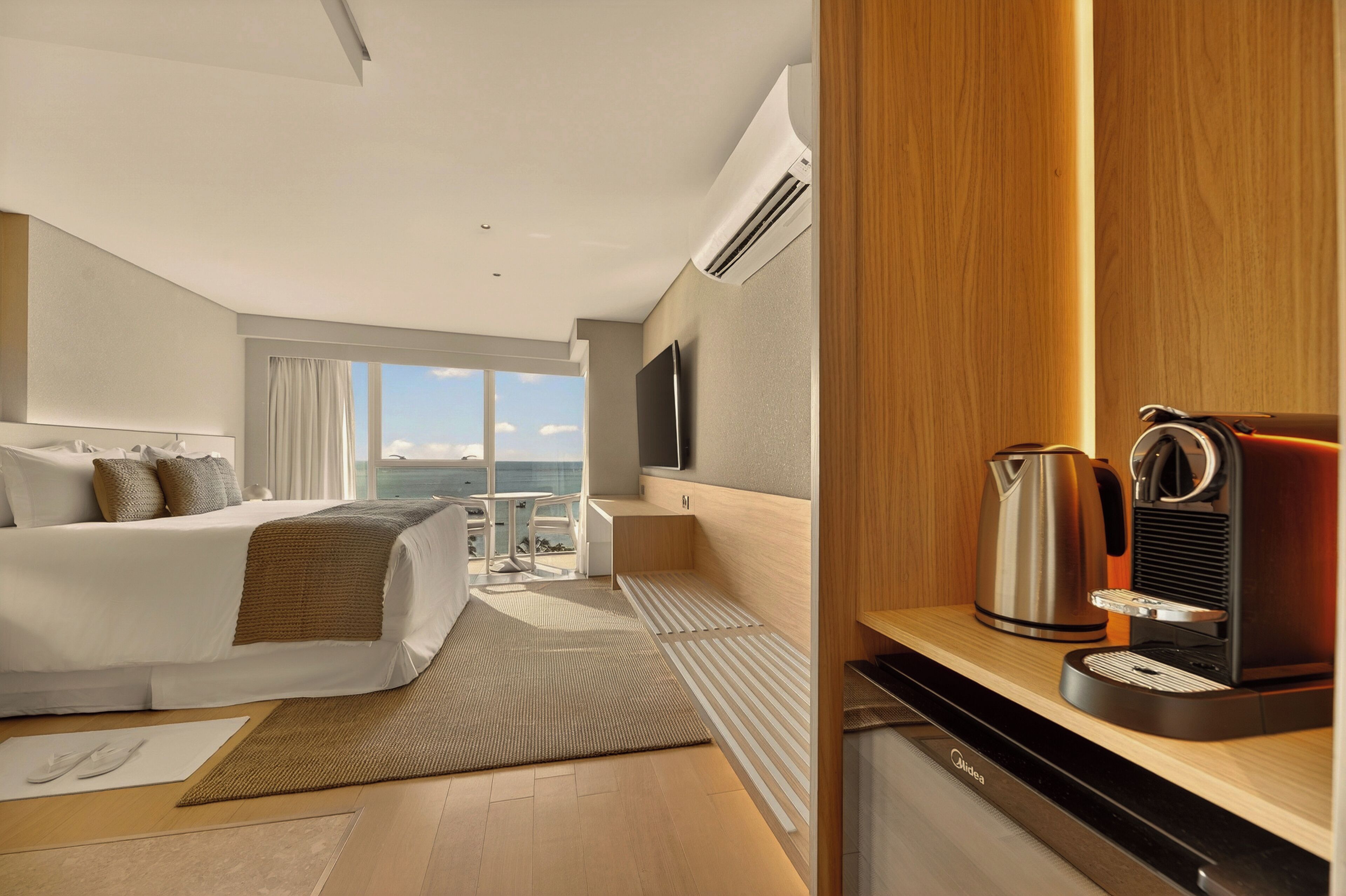 A modern hotel room with a bed, a TV, a coffee maker, and a view of the sea.