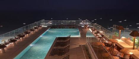 Une piscine sur le toit, agrémentée de chaises longues et de parasols, le tout entouré d’une rambarde en verre.