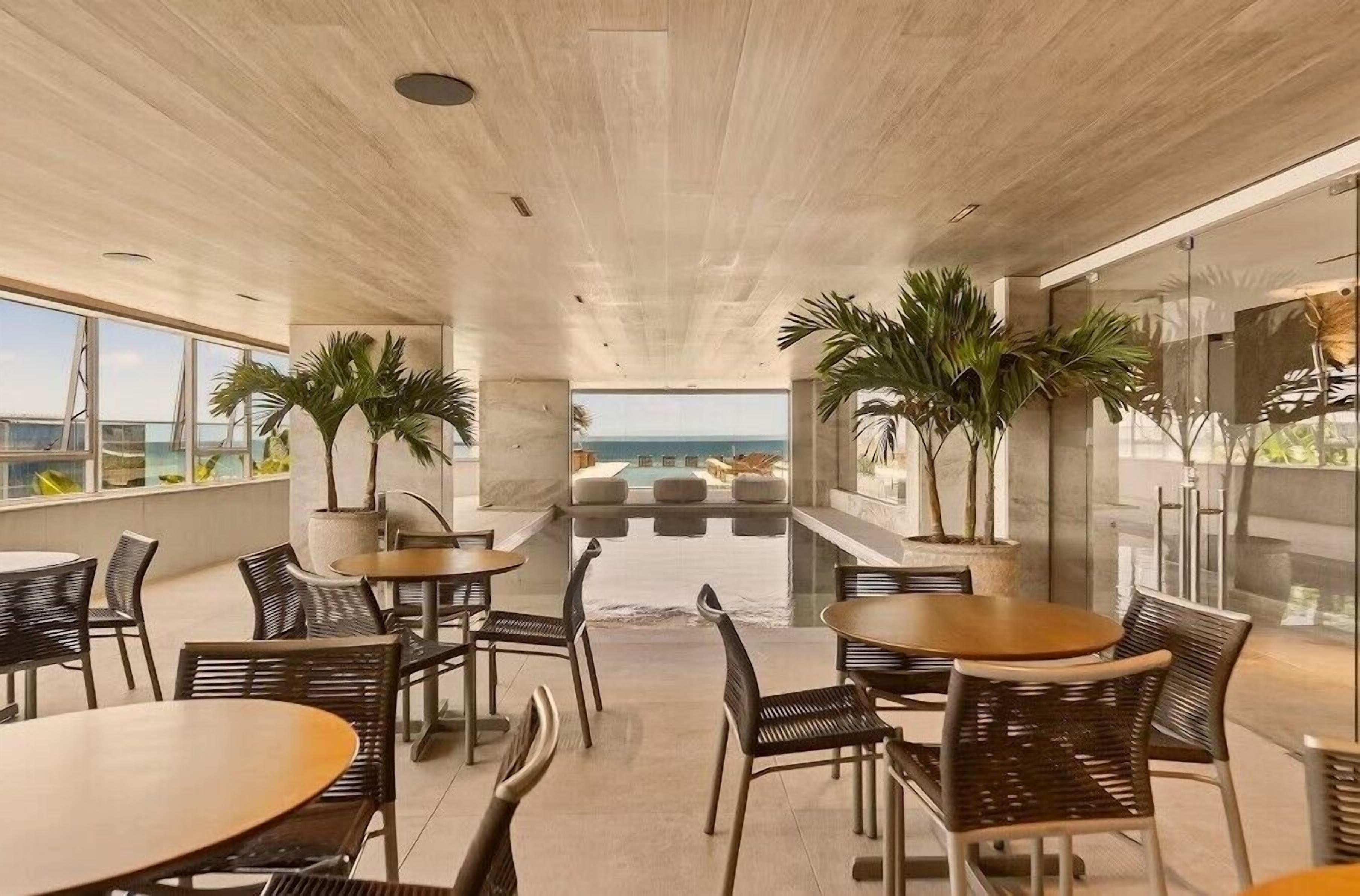 An indoor dining area with tables and chairs, large windows, and potted plants.