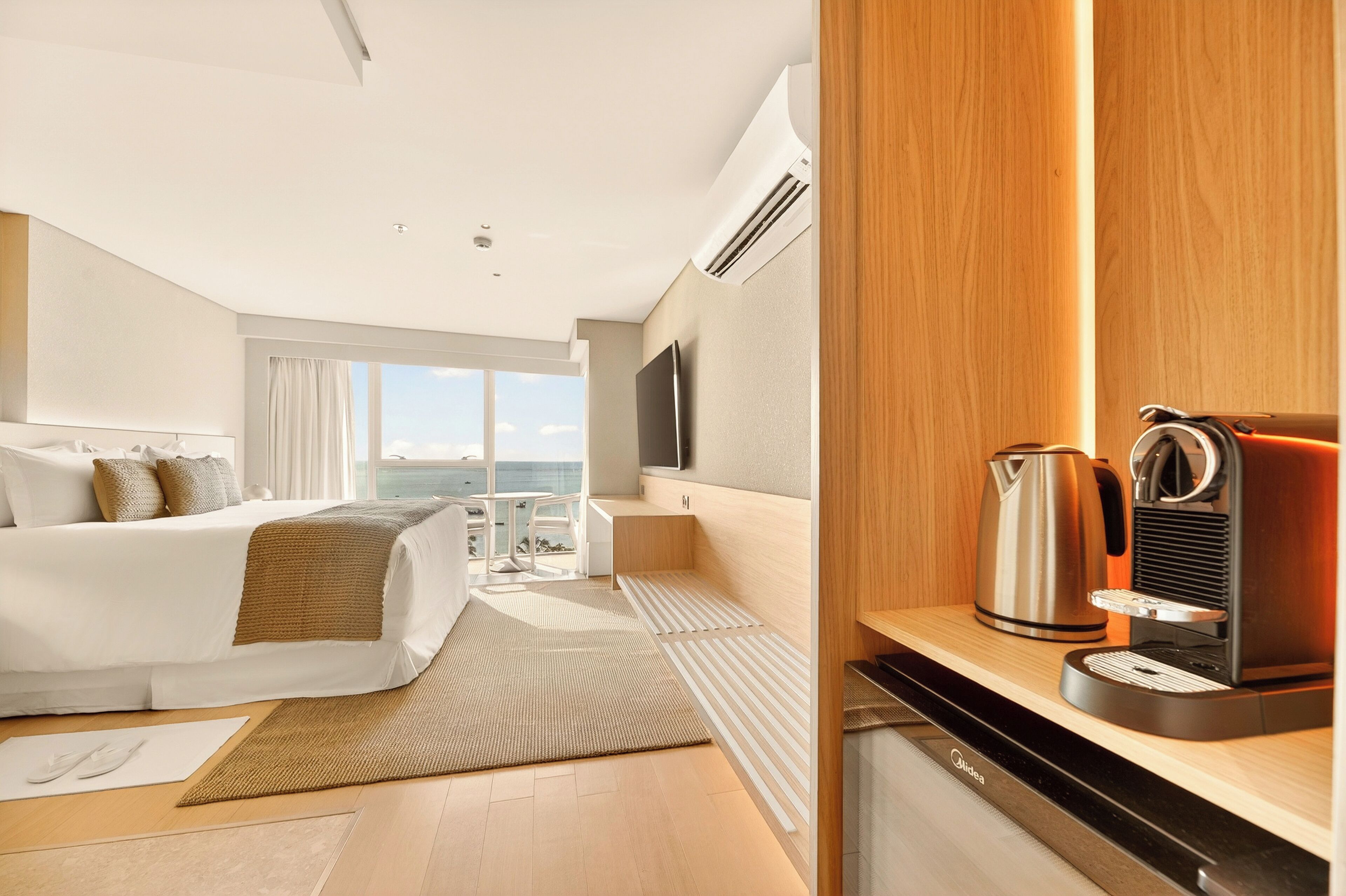 A modern hotel room with a bed, a desk with a coffee maker, a TV, and a view of the sea.