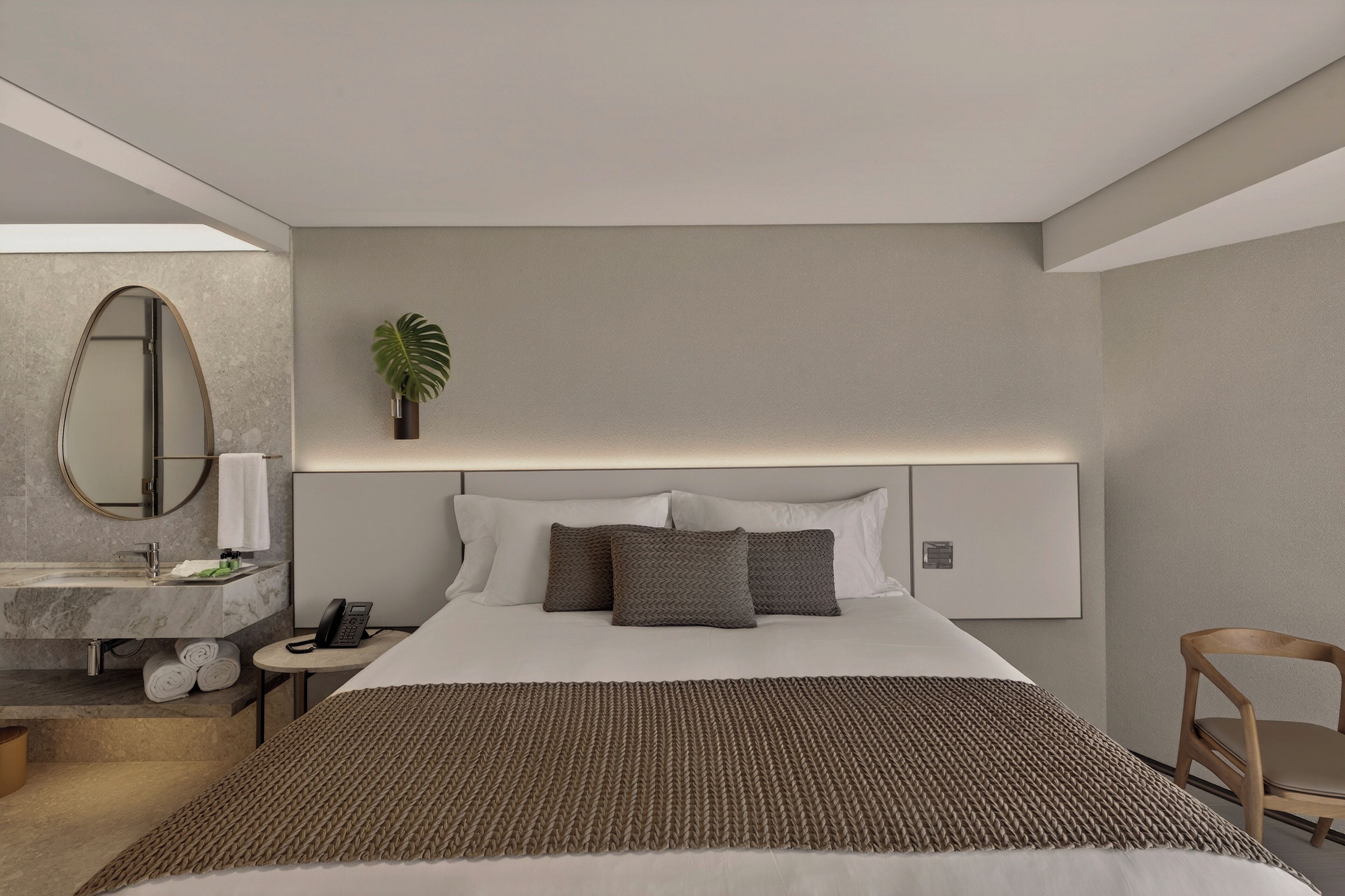 Habitación de hotel moderna con una cama grande, una mesita de noche con lámpara, un espejo, una planta y una silla de madera.
