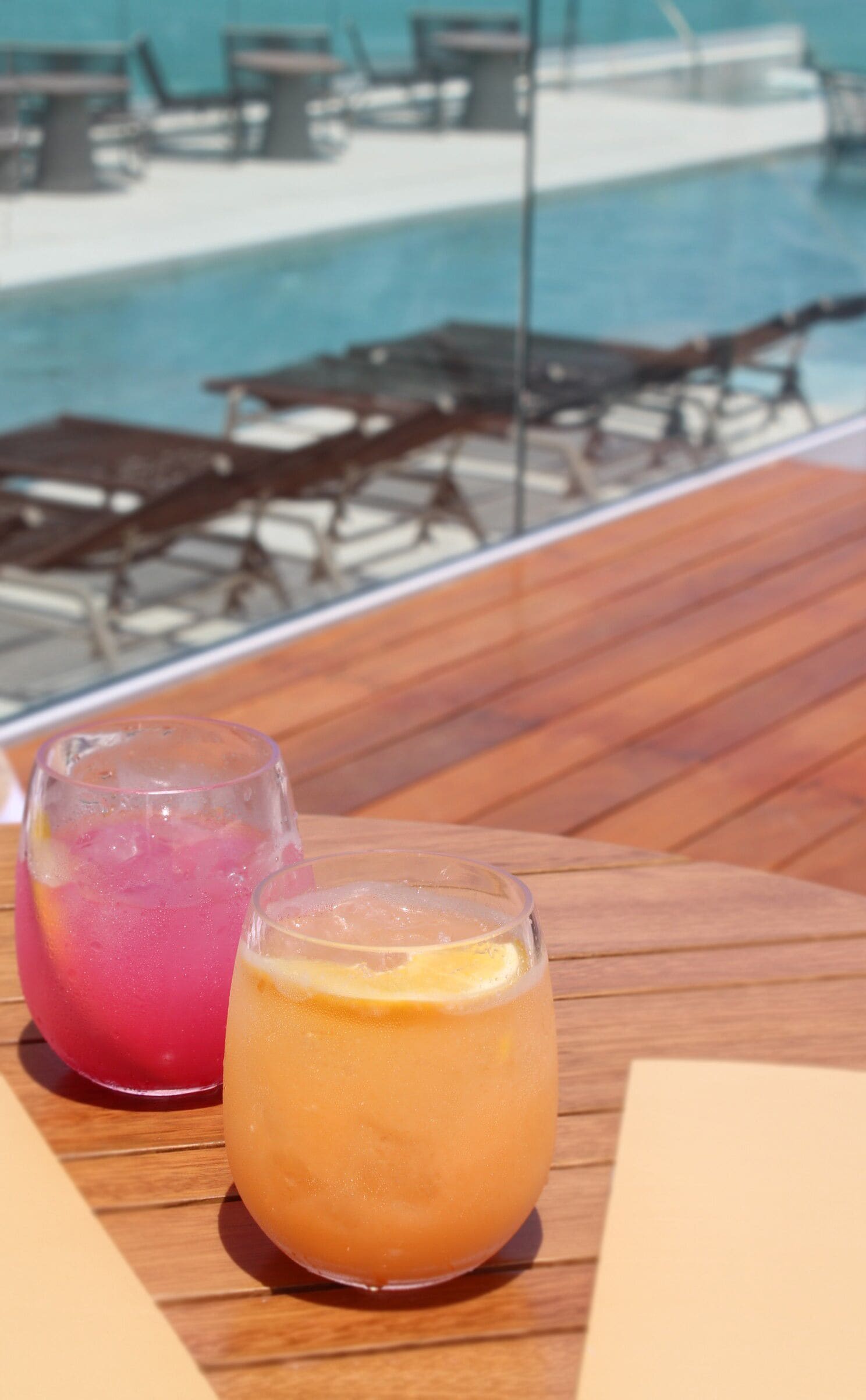 Two drinks on a wooden table with a pool and lounge chairs in the background.