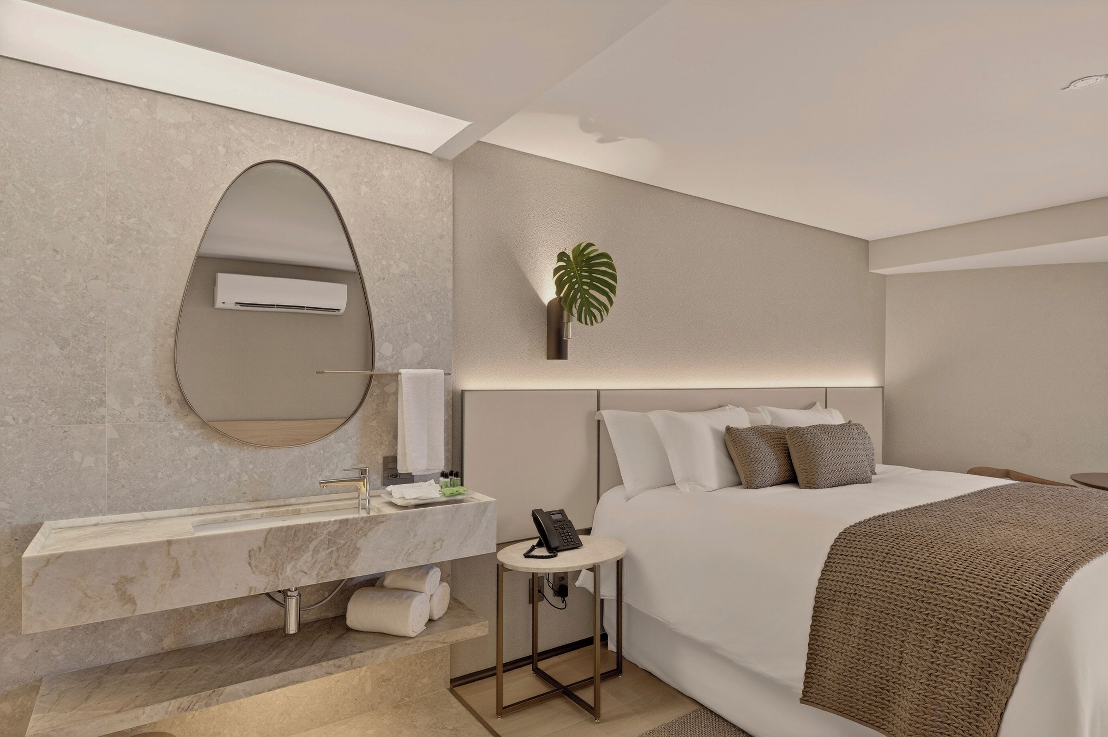 Una habitación de hotel moderna con una cama grande, un tocador empotrado con espejo, una mesita de noche pequeña con teléfono y una planta en maceta.