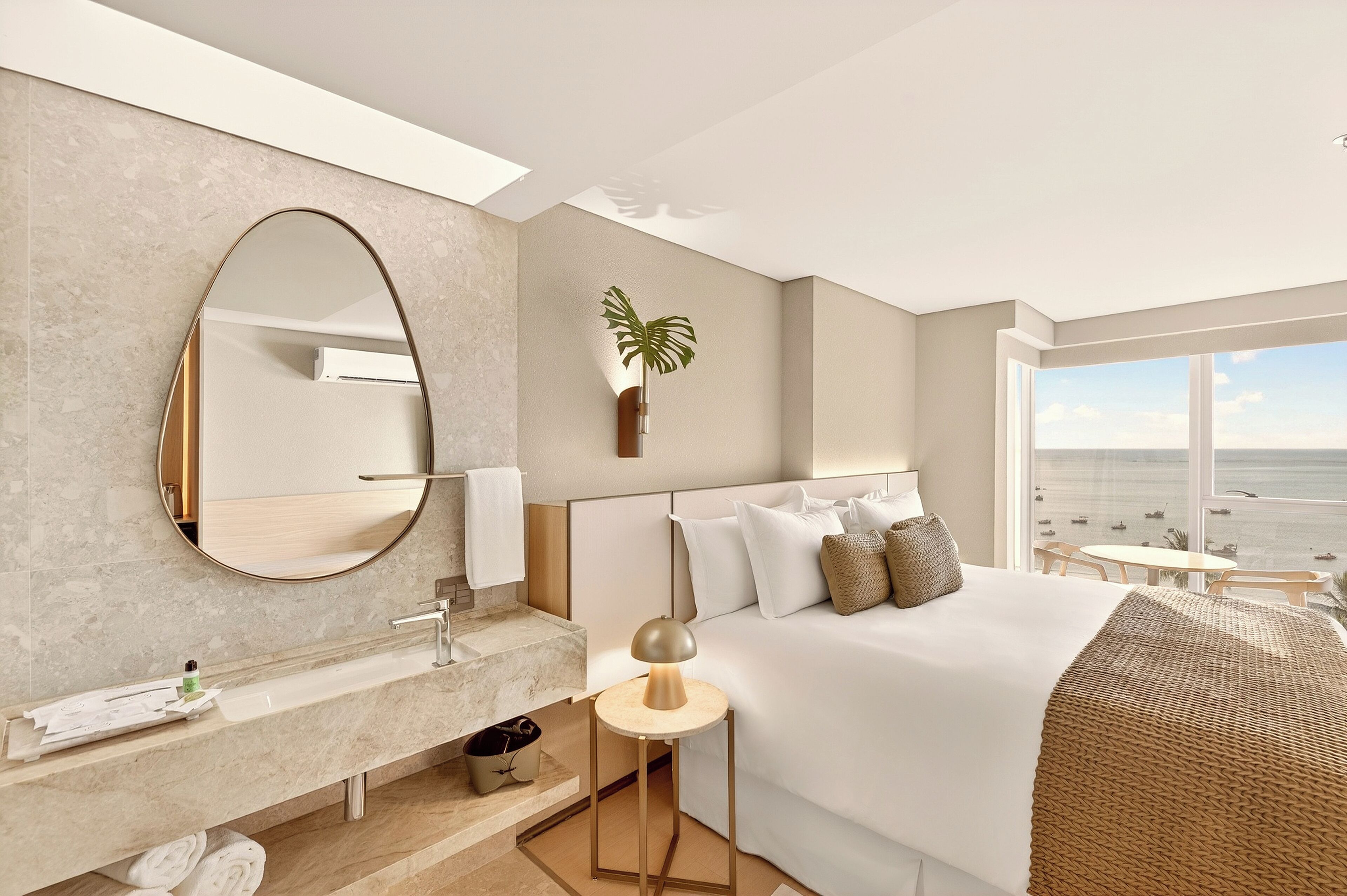 A modern hotel room with a large bed, a round mirror, a bedside lamp, a plant, and a view of the beach.