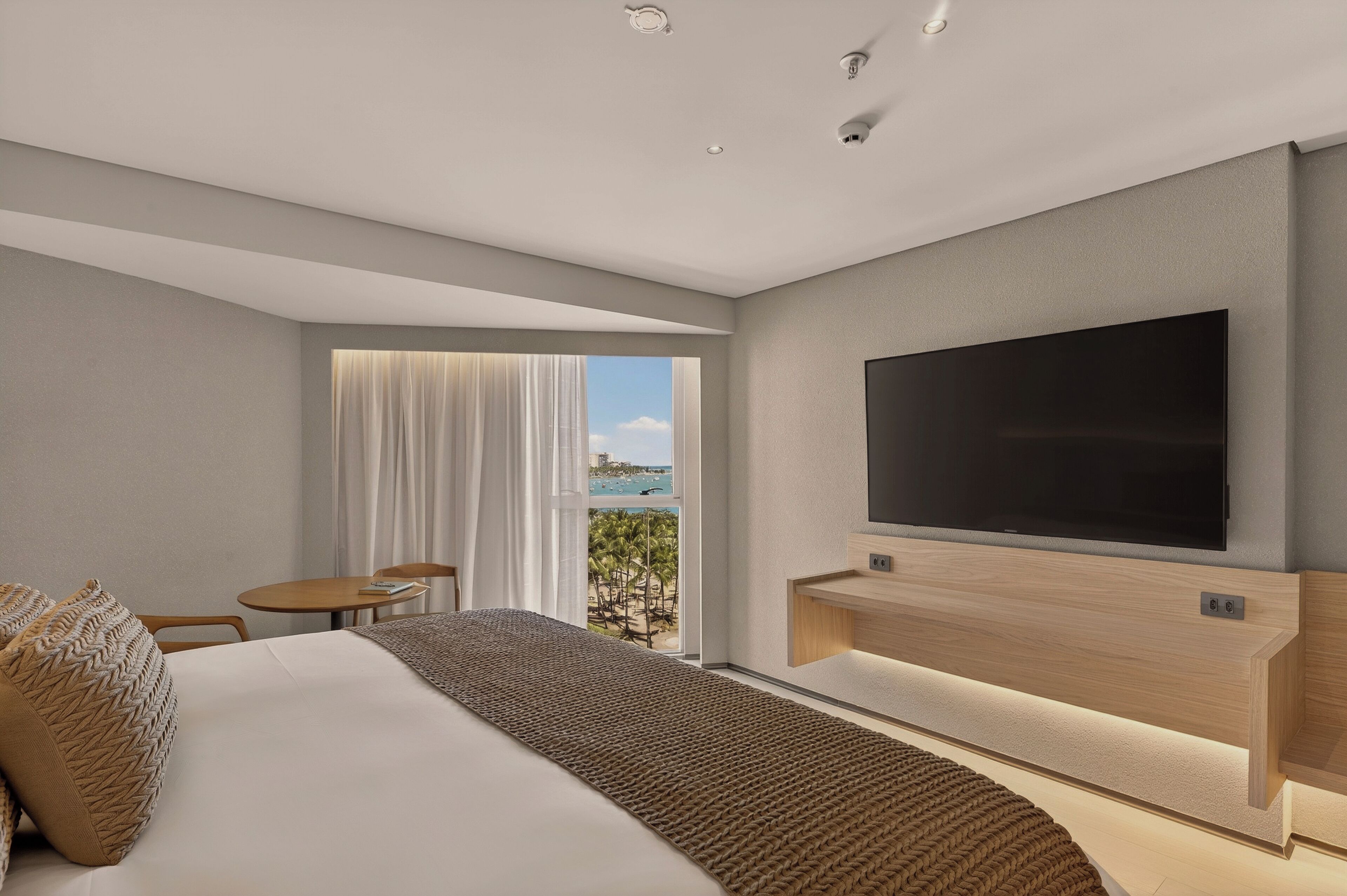A modern hotel room with a large bed, a flat-screen TV mounted on the wall, a small table and chair, and a window with a view of palm trees and the ocean.