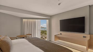 A modern hotel room with a large bed, a flat-screen TV mounted on the wall, a small table and chair, and a window with a view of palm trees and the ocean.