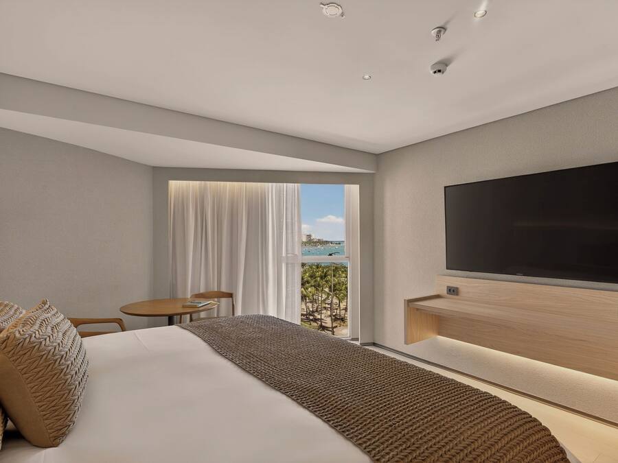 Een moderne hotelkamer met een groot bed, een flatscreen televisie aan de muur, een kleine tafel en stoel, en een raam met uitzicht op palmbomen en de oceaan.