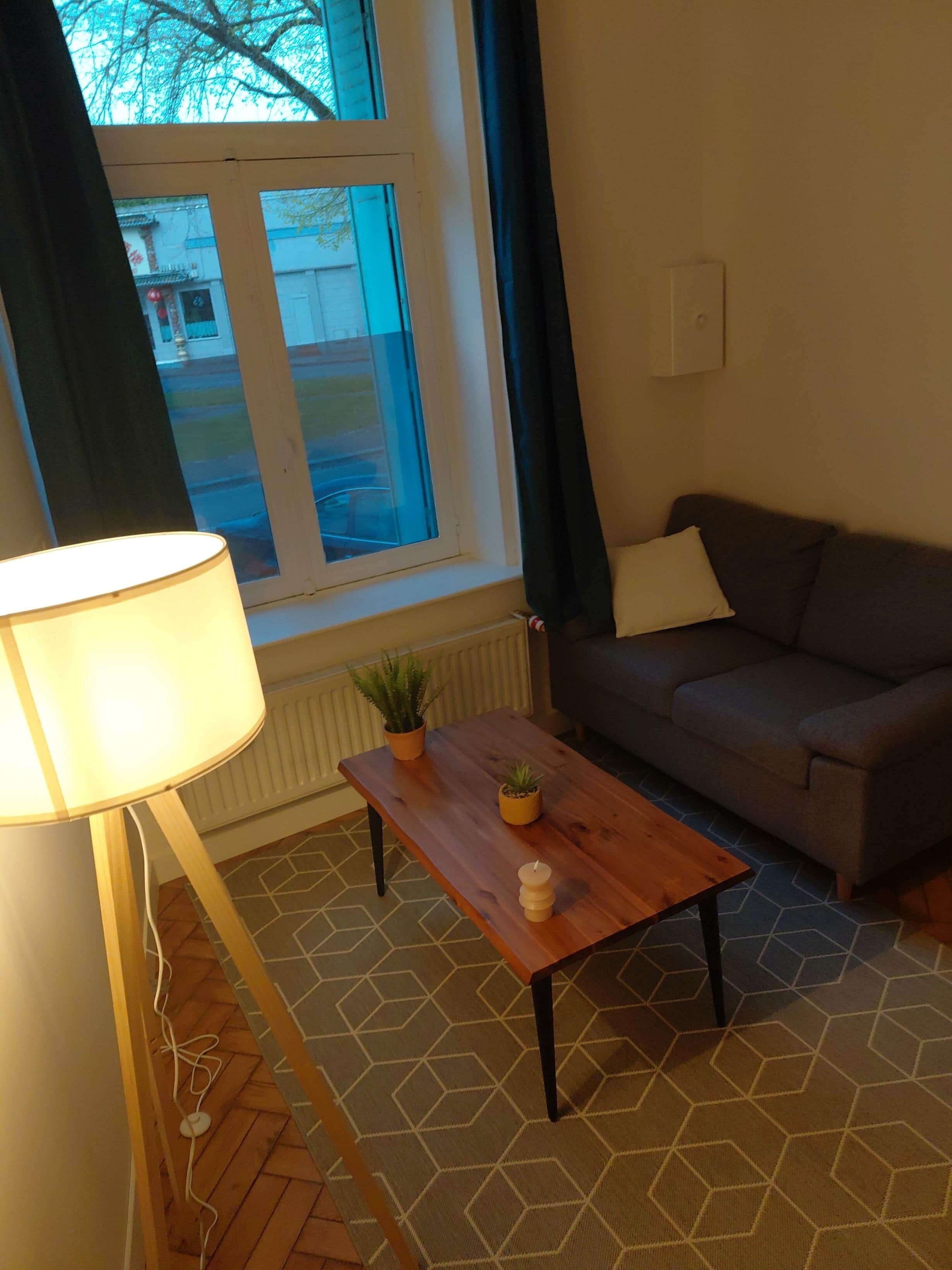 Ett rum med en soffa, ett träbord med en växt, en lampa och ett fönster med gardiner.