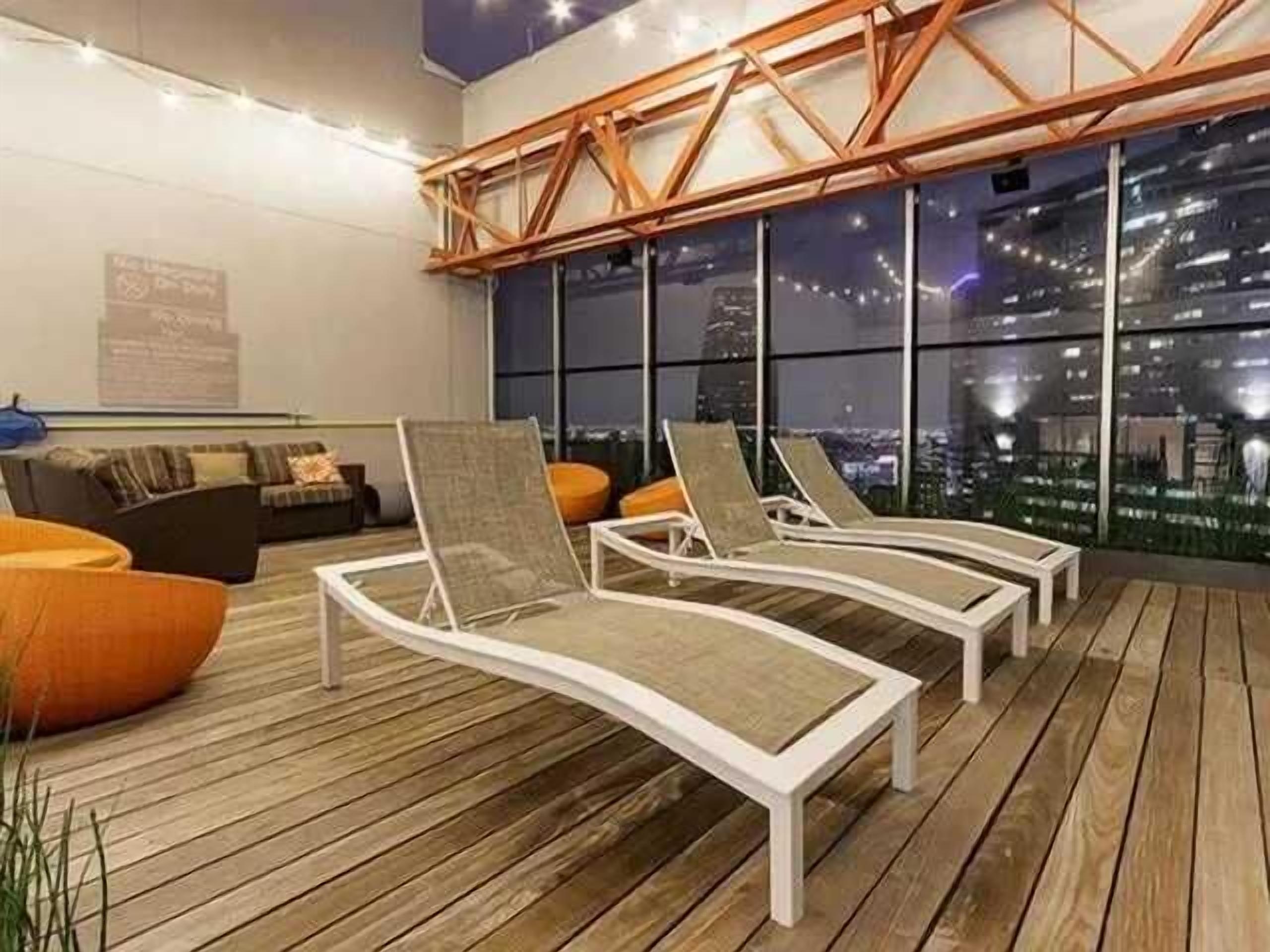Relax on modern rooftop loungers with panoramic city views and ambient string lighting. This stylish shared deck offers a peaceful setting to unwind, socialize, or enjoy the skyline in a comfortable outdoor space.