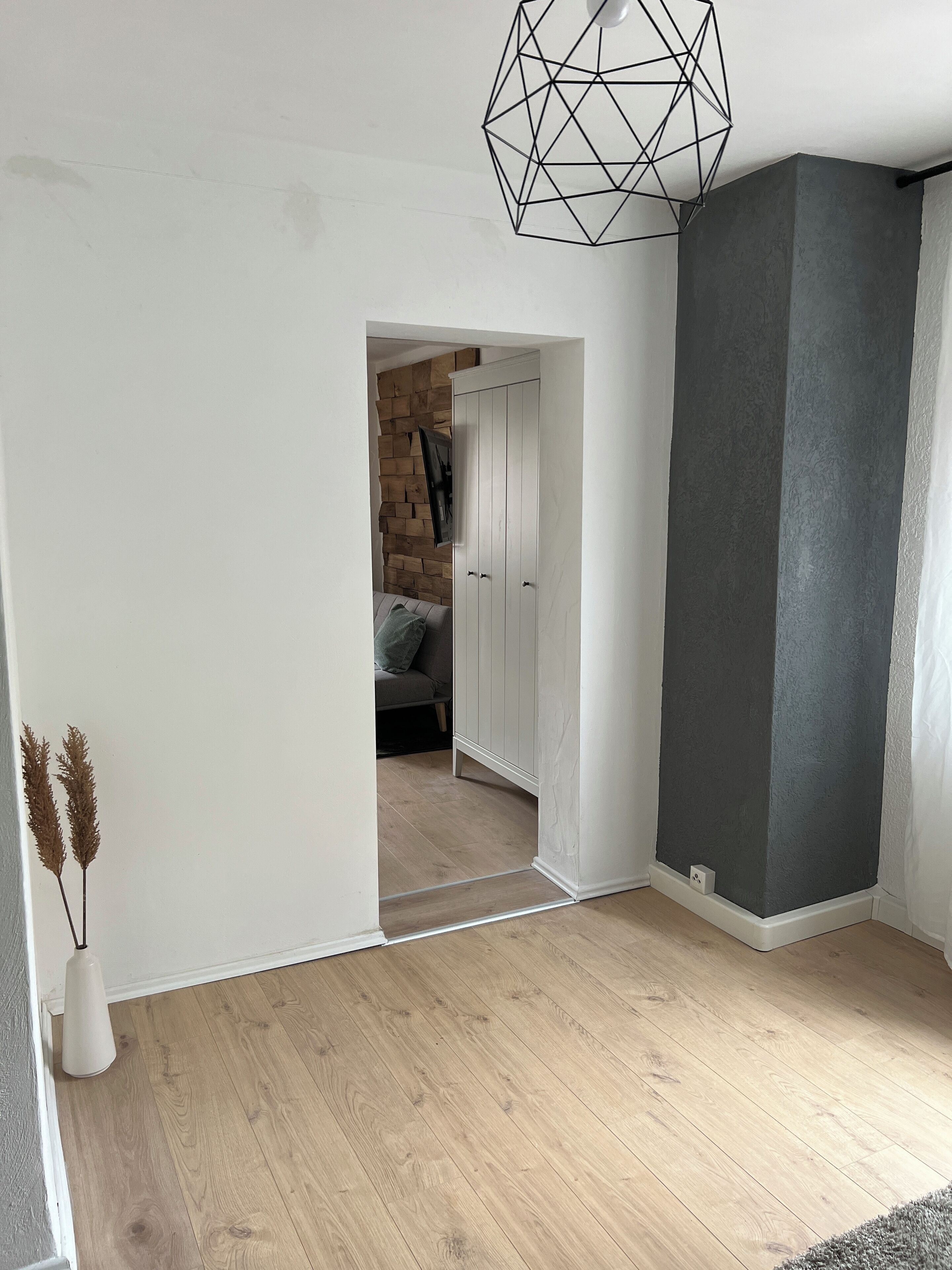 Ein Zimmer mit Holzboden, einer weißen Tür, die in einen anderen Raum führt, und einer geometrisch gestalteten Leuchte.
