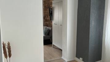Ein Zimmer mit Holzboden, einer weißen Tür, die in einen anderen Raum führt, und einer geometrisch gestalteten Leuchte.
