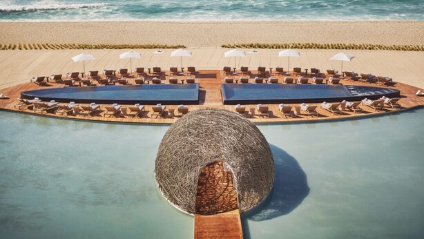 Un espace aménagé au bord de la piscine, avec des chaises longues et des parasols, entouré d’une plage de sable et de l’océan.