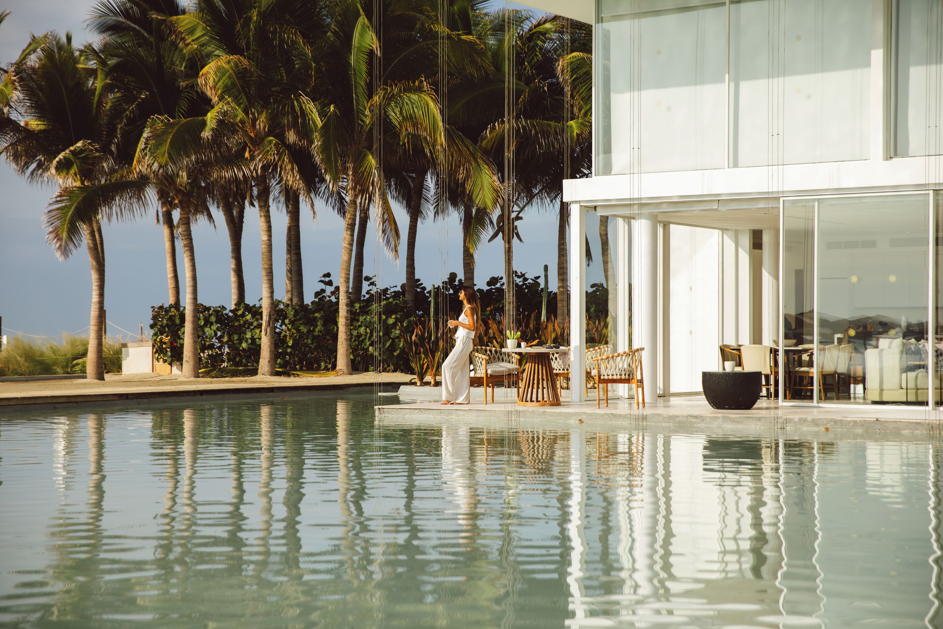 Une piscine bordée de palmiers, un bâtiment moderne aux grandes fenêtres et du mobilier d’extérieur.