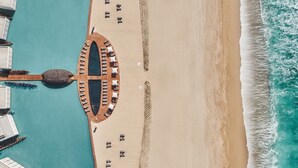 Vue aérienne d’un complexe hôtelier situé en bord de mer, avec une piscine, des chaises longues et une terrasse en bois.