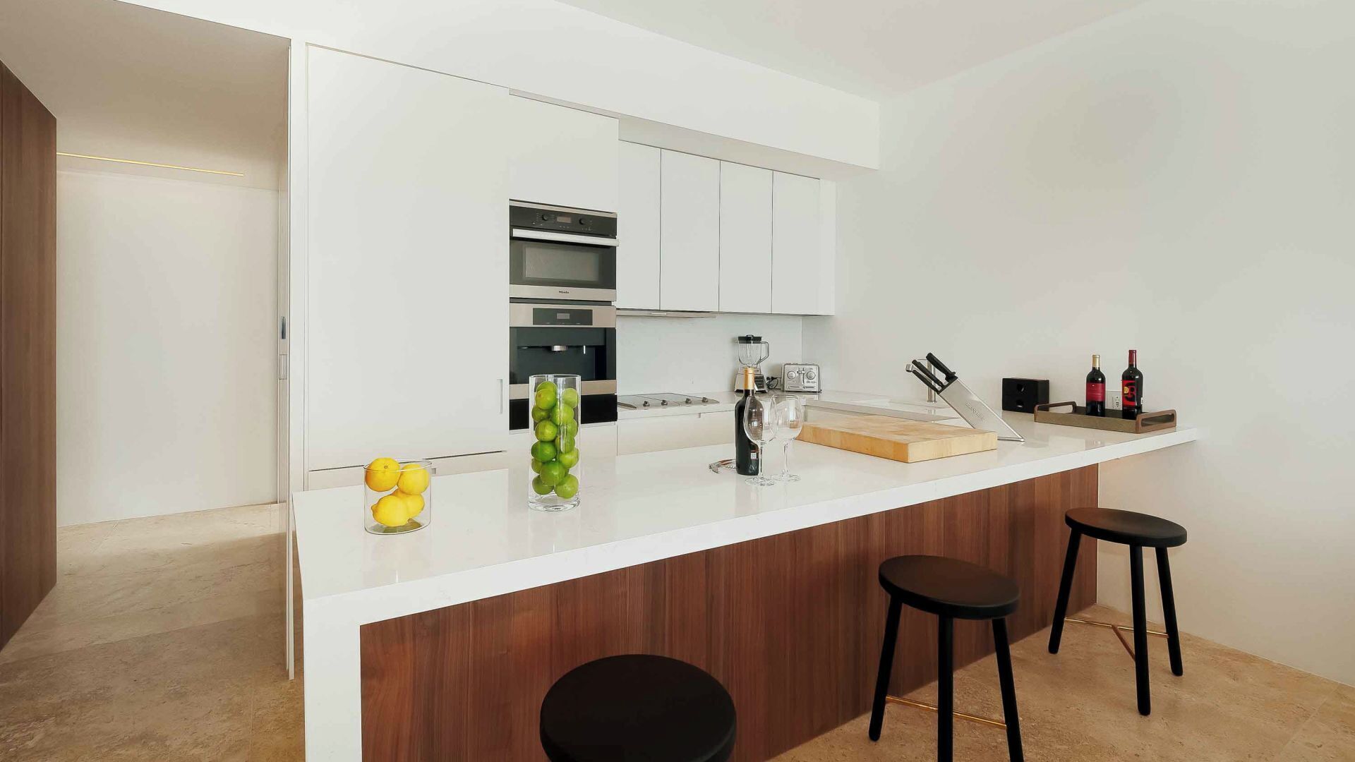 A modern kitchen with white cabinets, a built-in oven, a wine bottle, and two stools.