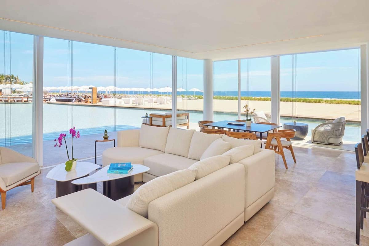 Un vaste espace de vie avec une grande fenêtre donnant sur une plage et une piscine. On y trouve un canapé, une table à manger et une petite table basse agrémentée de livres et de fleurs.