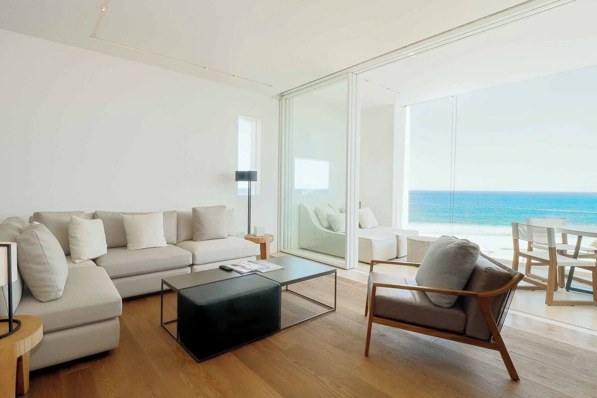 Un salon moderne avec un canapé, une table basse et un fauteuil, un lampadaire et une vue sur l’océan par de grandes fenêtres.