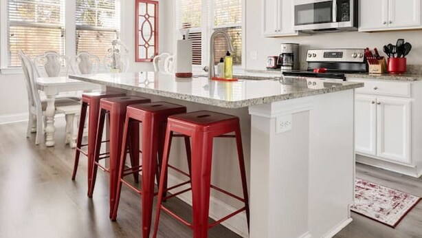Uma cozinha moderna com armários brancos, uma ilha central com banquetas vermelhas, eletrodomésticos de aço inoxidável e iluminação pendente.