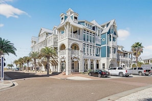 A large, multi-story building with a prominent balcony, a stone facade, and a clear blue sky. - Lala Land TC 6302 (Port Aransas)