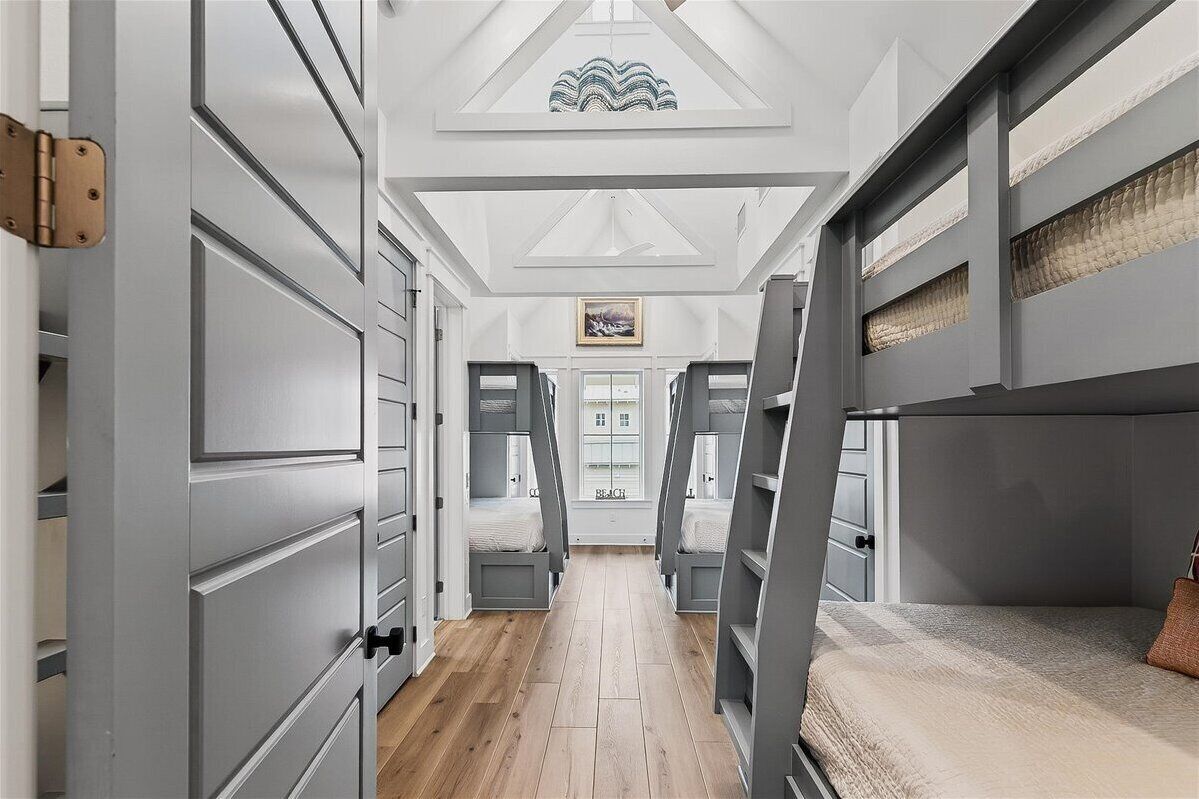 A narrow hallway with a high ceiling, wooden flooring, and grey walls. There are two bunk beds on either side of the hallway, and a door at the end with a framed picture above it.
