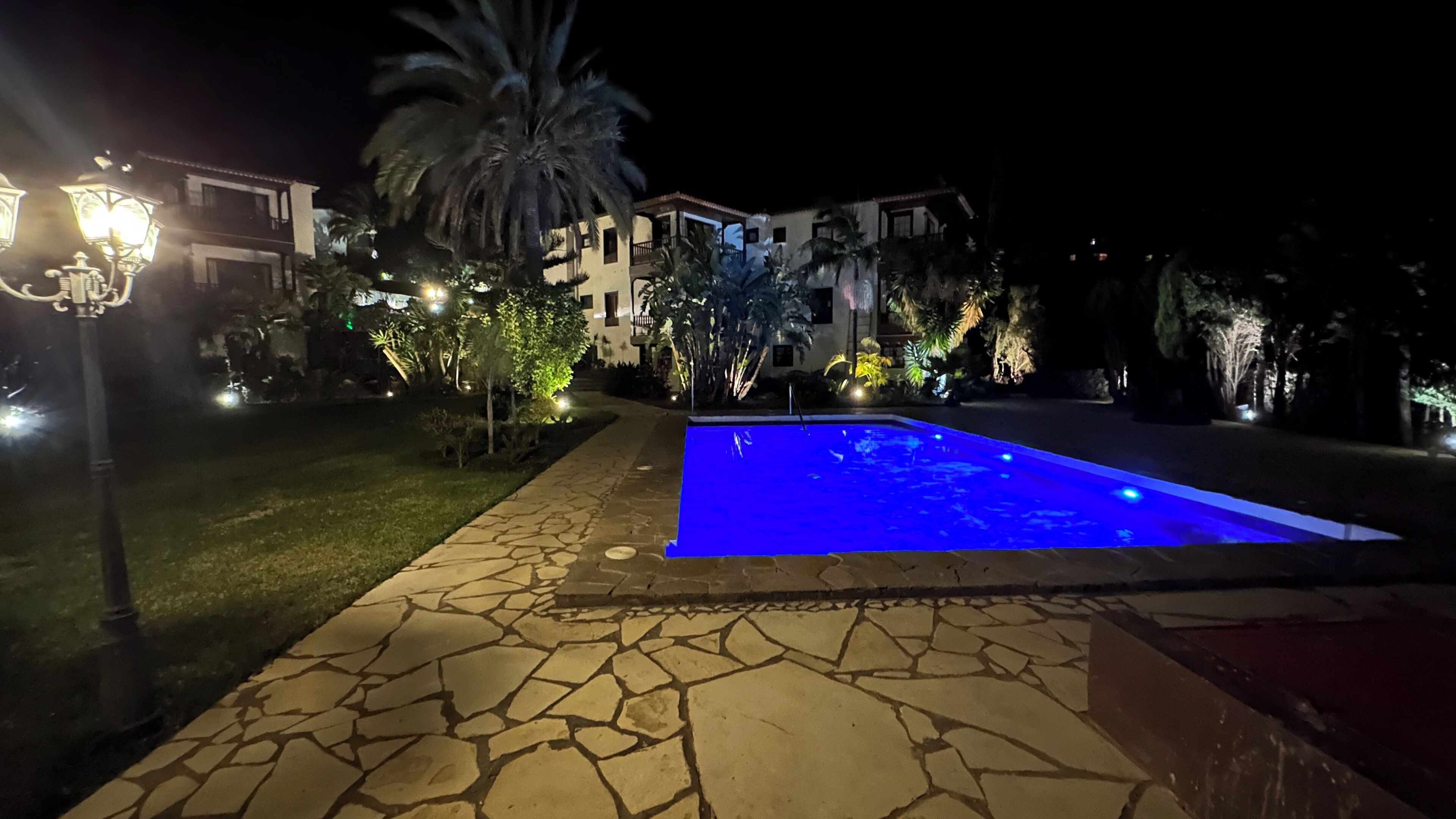 A nighttime view of a pool area with stone flooring and a blue-lit pool, surrounded by well-lit buildings and palm trees.