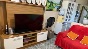 Un salon avec un grand téléviseur à écran plat posé sur un meuble en bois, un canapé rouge avec des coussins orange, un ventilateur et une petite table sur laquelle se trouvent une bouteille et une photo encadrée.