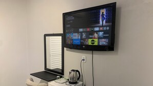 Een compacte kamer met een aan de muur bevestigde flatscreen-tv waarop diverse streamingdiensten worden weergegeven. Een kleine, witte koelkast staat tegen de muur en er is een raam met gesloten jaloezieën zichtbaar.