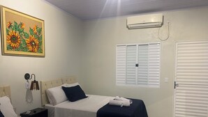 Une chambre d’hôtel avec deux lits, un tableau représentant des tournesols, un climatiseur, une fenêtre avec des stores et une porte.