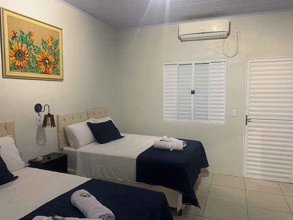A hotel room with two beds, a painting of sunflowers, an air conditioner, a window with blinds, and a door.