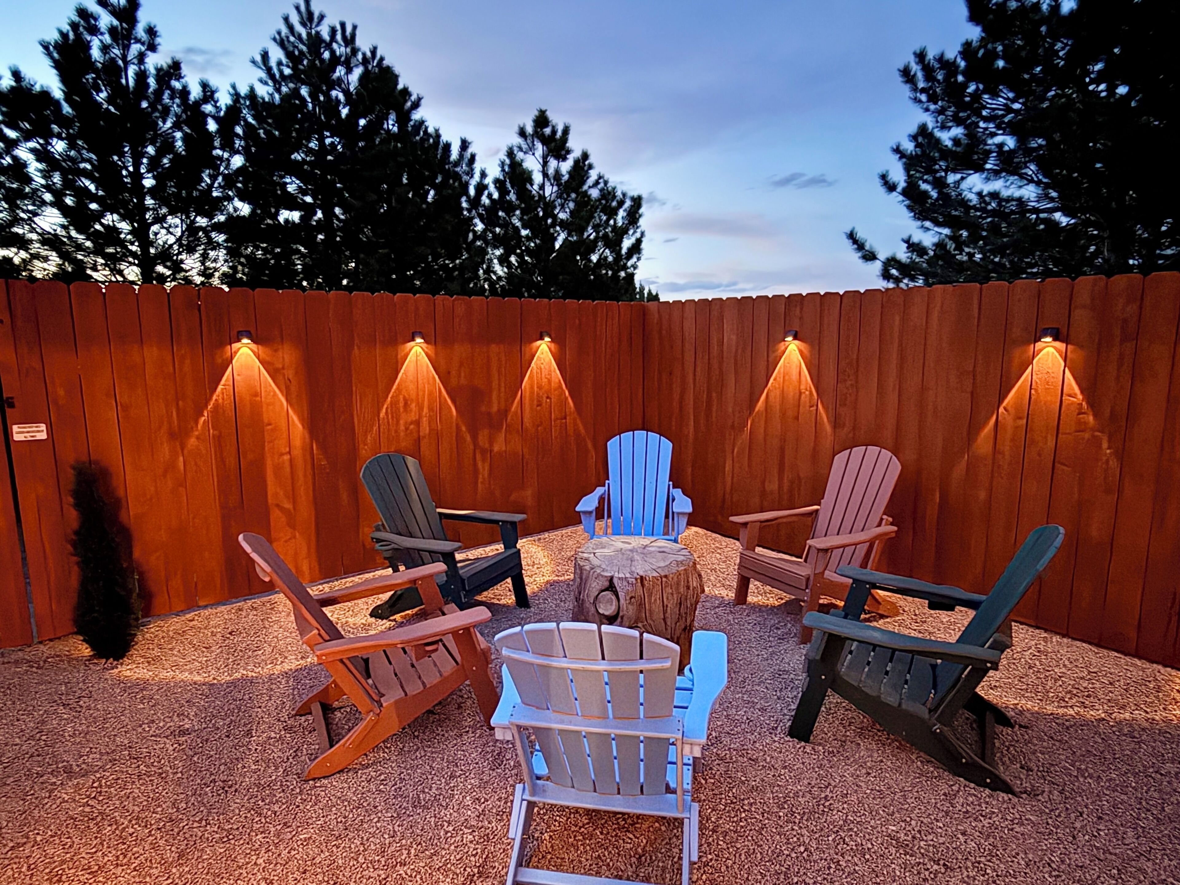 Eine Terrasse mit Adirondack-Stühlen und einem Feuerkorb in der Mitte, umgeben von einem Holzzaun und Bäumen.