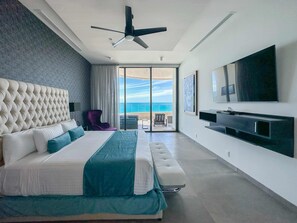 Un dormitorio moderno con una cama grande, ventilador de techo, televisión y balcón con vista al océano.