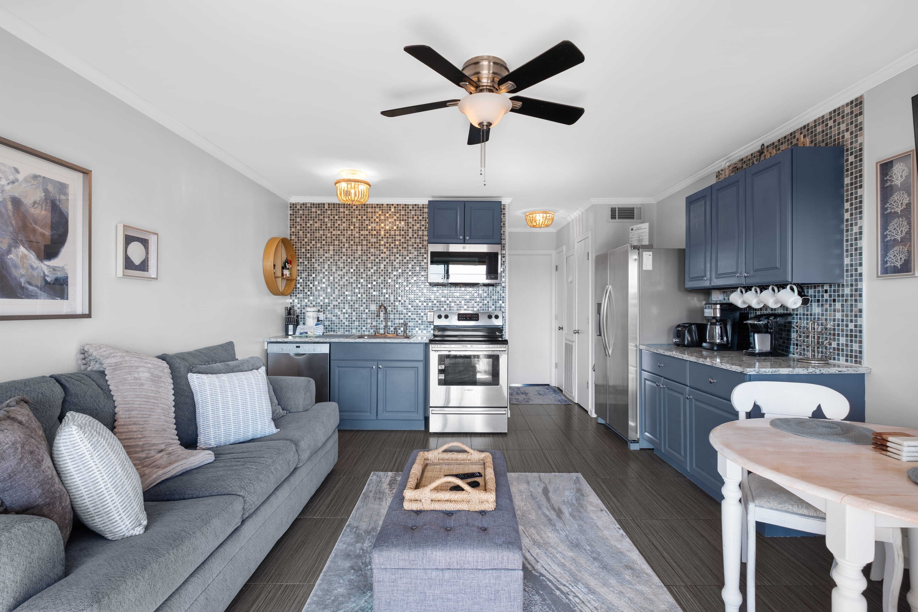 Una cocina moderna con armarios azul oscuro, un refrigerador de acero inoxidable y un microondas. Hay un sofá gris con cojines estampados y una mesa de centro de madera.