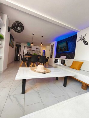 Uma sala de estar moderna com um sofá branco, uma mesa de jantar com cadeiras e uma cozinha ao fundo.