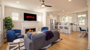Ein modernes Wohnzimmer mit Kamin, einem an der Wand montierten Fernseher und einer Küche im Hintergrund.