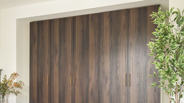 Une chambre avec une grande armoire, un lit recouvert d’une courtepointe blanche et un tapis à motifs.