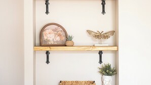Une étagère murale en bois, composée de trois étagères, sur lesquelles sont disposés des objets décoratifs, comme un tableau encadré, une plante en pot et un panier tressé.