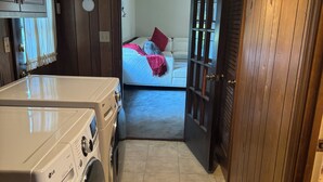 Ein Waschraum mit einer LG Waschmaschine und einem Trockner, einer Holztür zu einem Schlafzimmer mit Bett und Fenster.