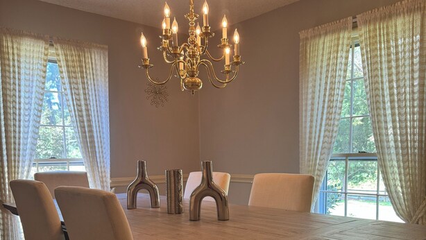 Ein Esszimmer mit einem großen Holztisch, sechs Stühlen, einer Chandelierleuchtre und großen Fenstern mit Vorhängen.