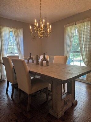 Een eetkamer met een grote houten tafel, zes stoelen, een kroonluchter en grote ramen voorzien van gordijnen.