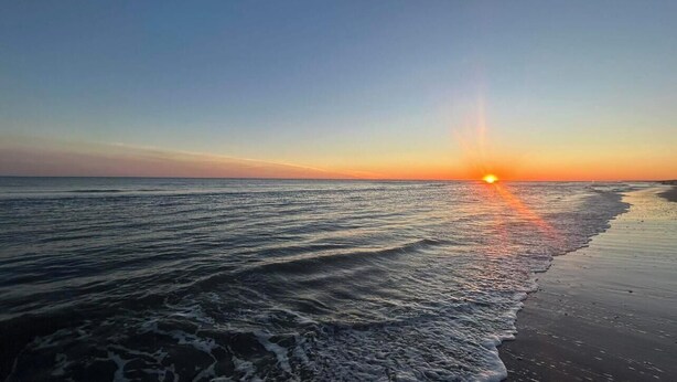 A beach at sunset with the sun low on the horizon, waves gently lapping the shore.