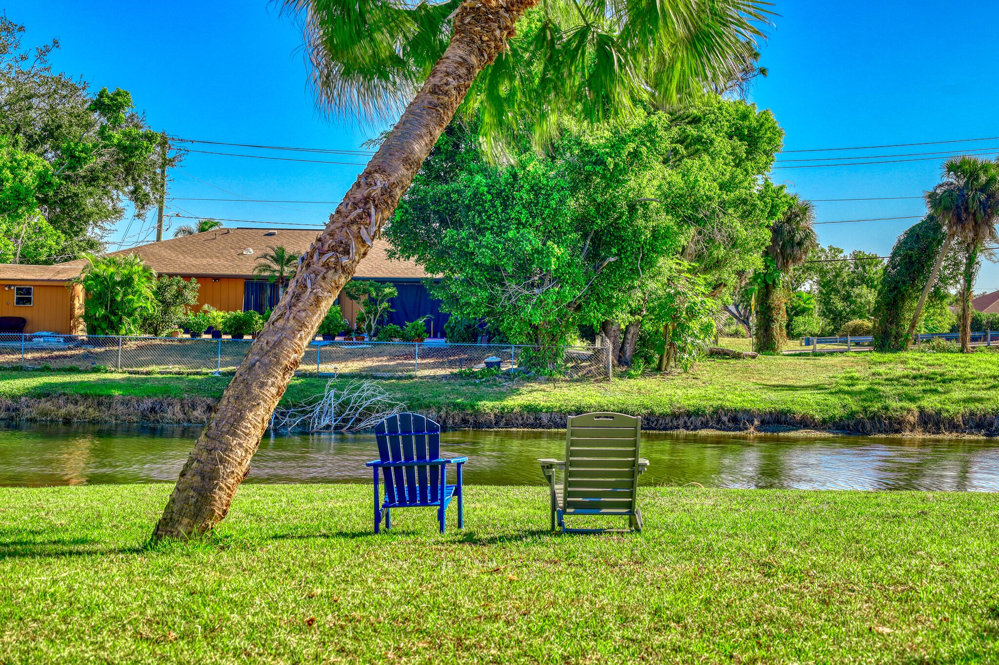 A serene riverside setting with two Adirondack chairs, a palm tree, and a house in the background.