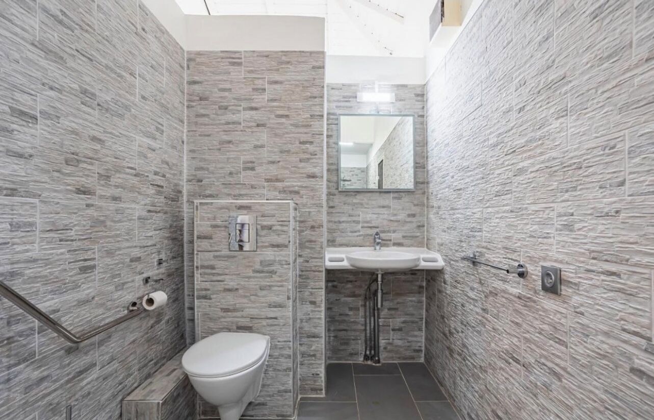 Et badeværelse med grå stenvægge, et hvidt toilet og en vask med et spejl over.