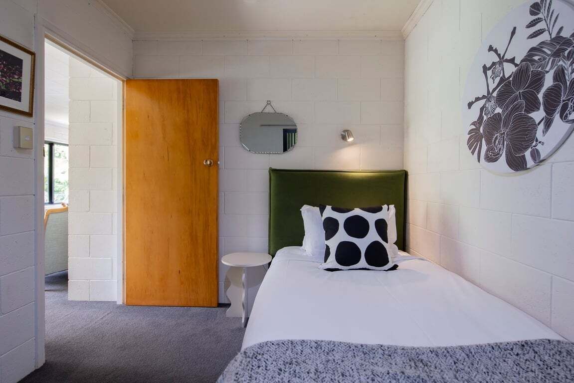 Een slaapkamer met een groen bekleden hoofdeinde, een witte spreidek en een ronde spiegel, plus een bloemendecoratie aan de muur.