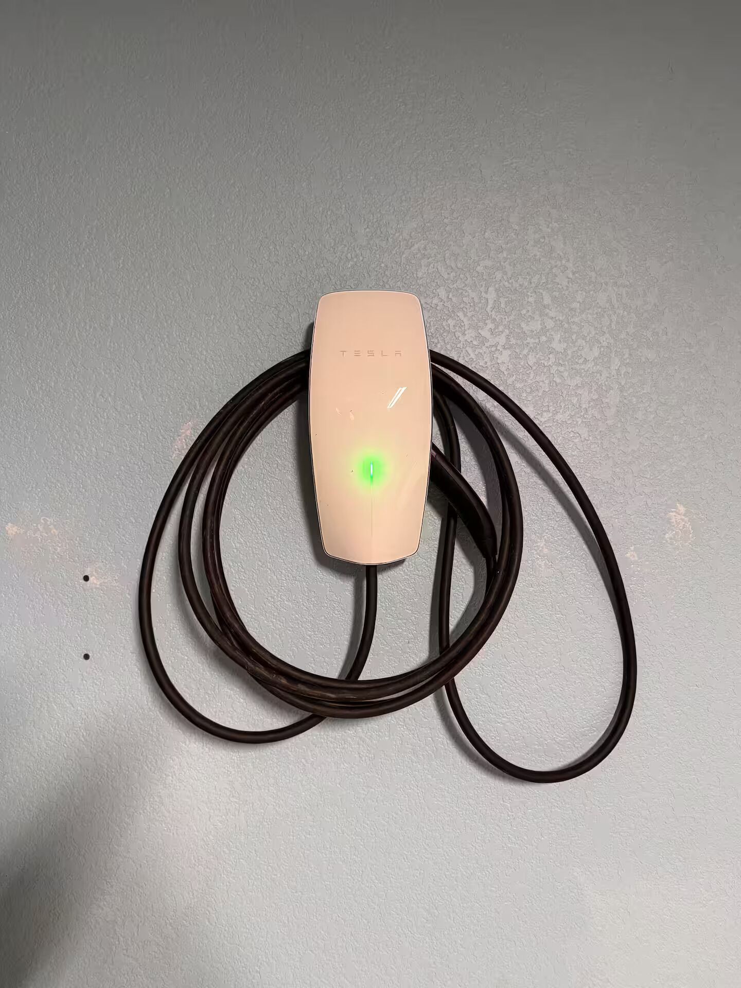 Une borne de recharge Tesla avec un voyant vert sur une surface grise texturée.