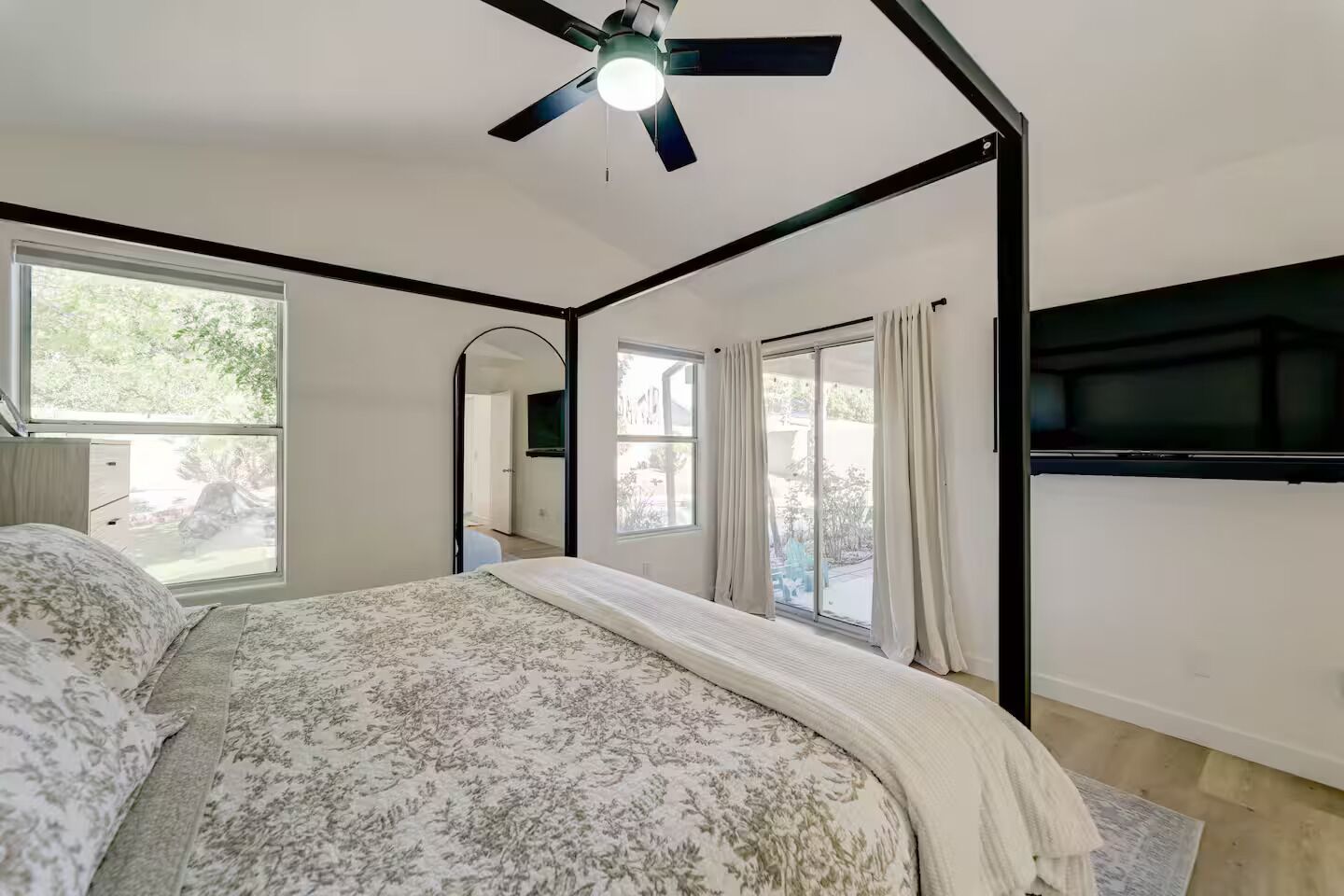 A bedroom with a bed, a ceiling fan, a TV mounted on the wall, and large windows with curtains.