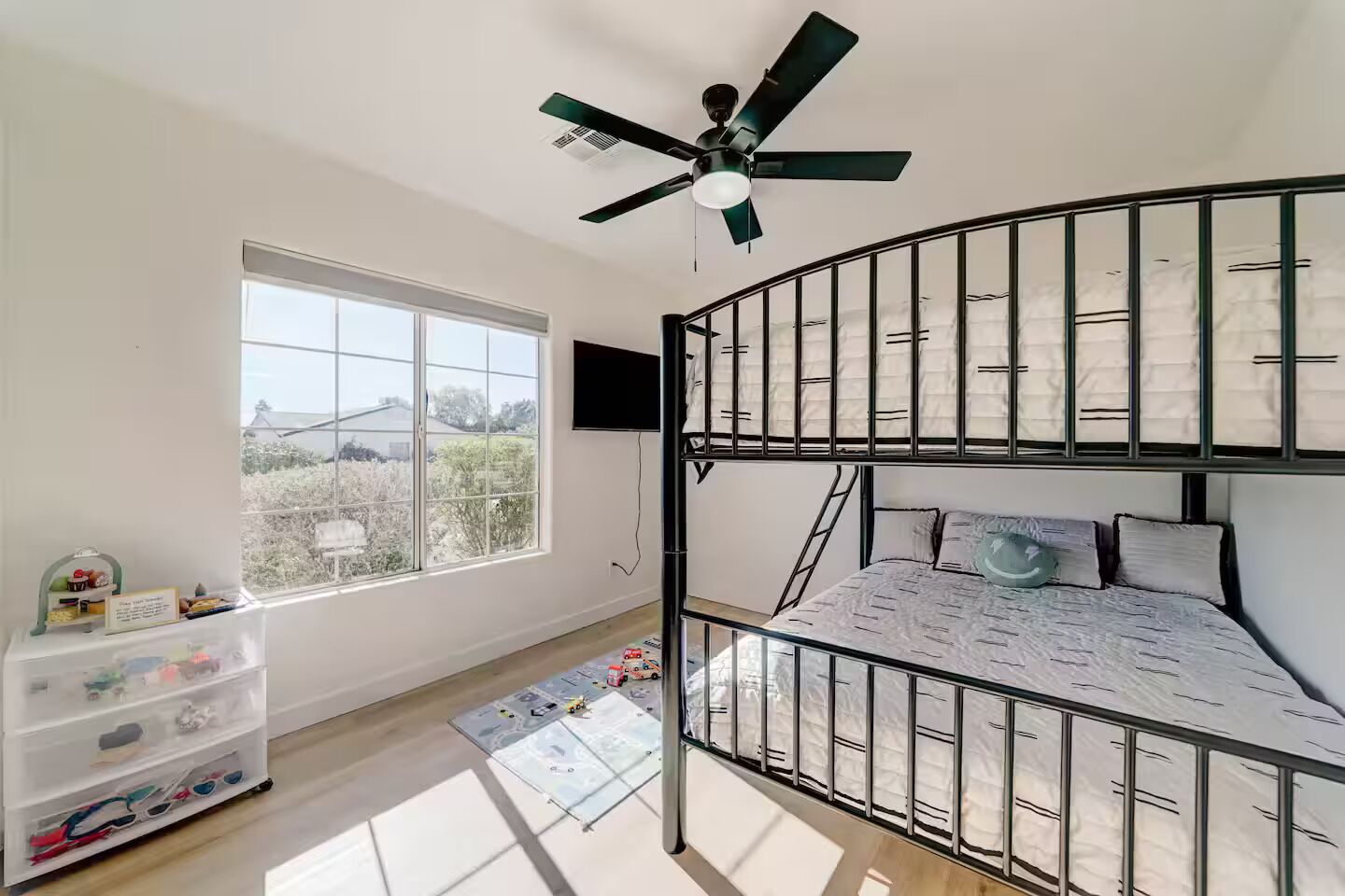 Een slaapkamer met stapelbed, een plafondventilator, een raam met uitzicht en een opbergmeubel met speelgoed.