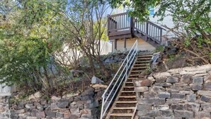 Escadas de pedra levam a um deck de madeira com guarda-corpo metálico, cercado por uma parede de pedra e vegetação.