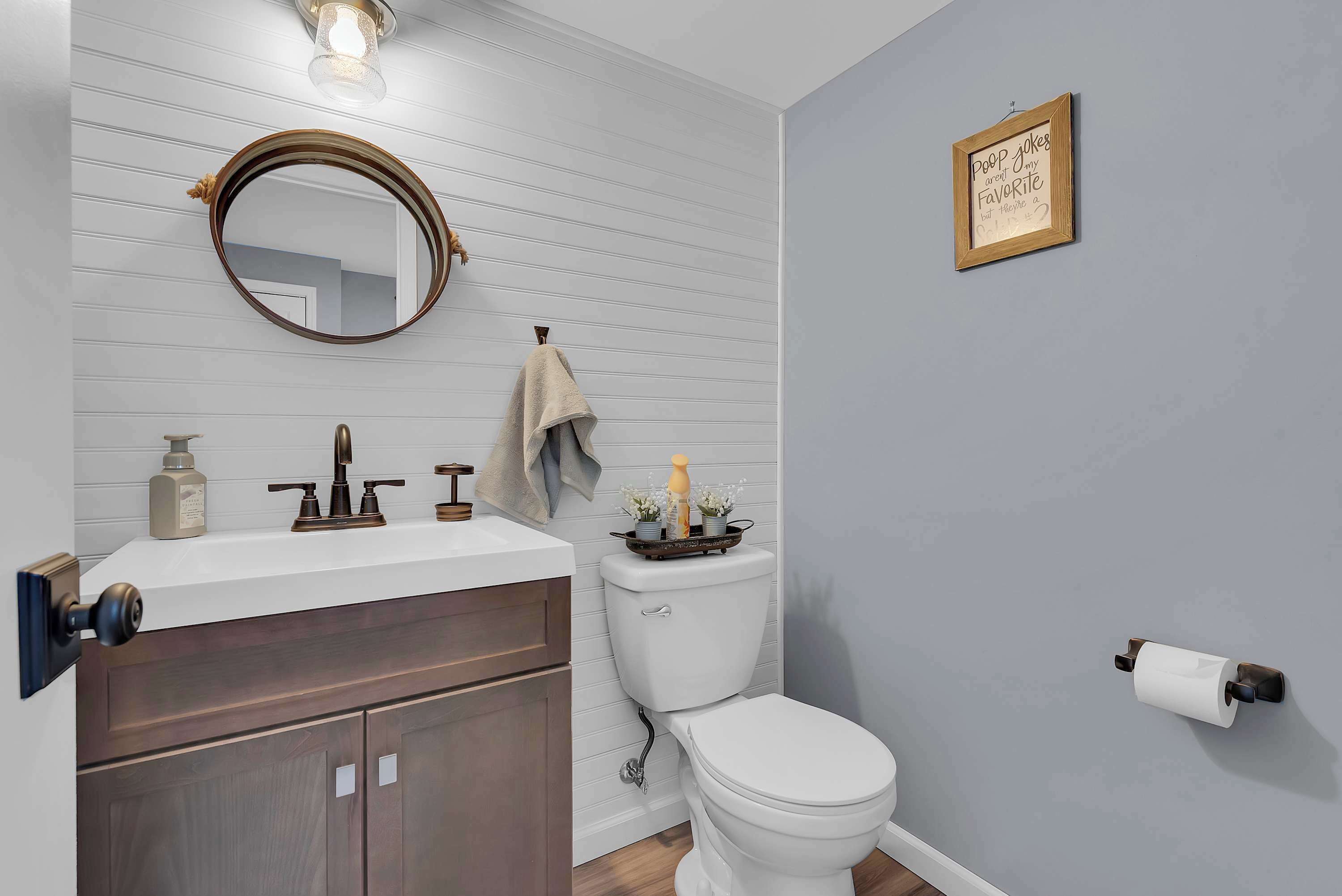 Une salle de bain avec un lavabo blanc, des meubles en bois foncé, un miroir rond, des toilettes et un porte-rouleau de papier toilette fixé au mur.