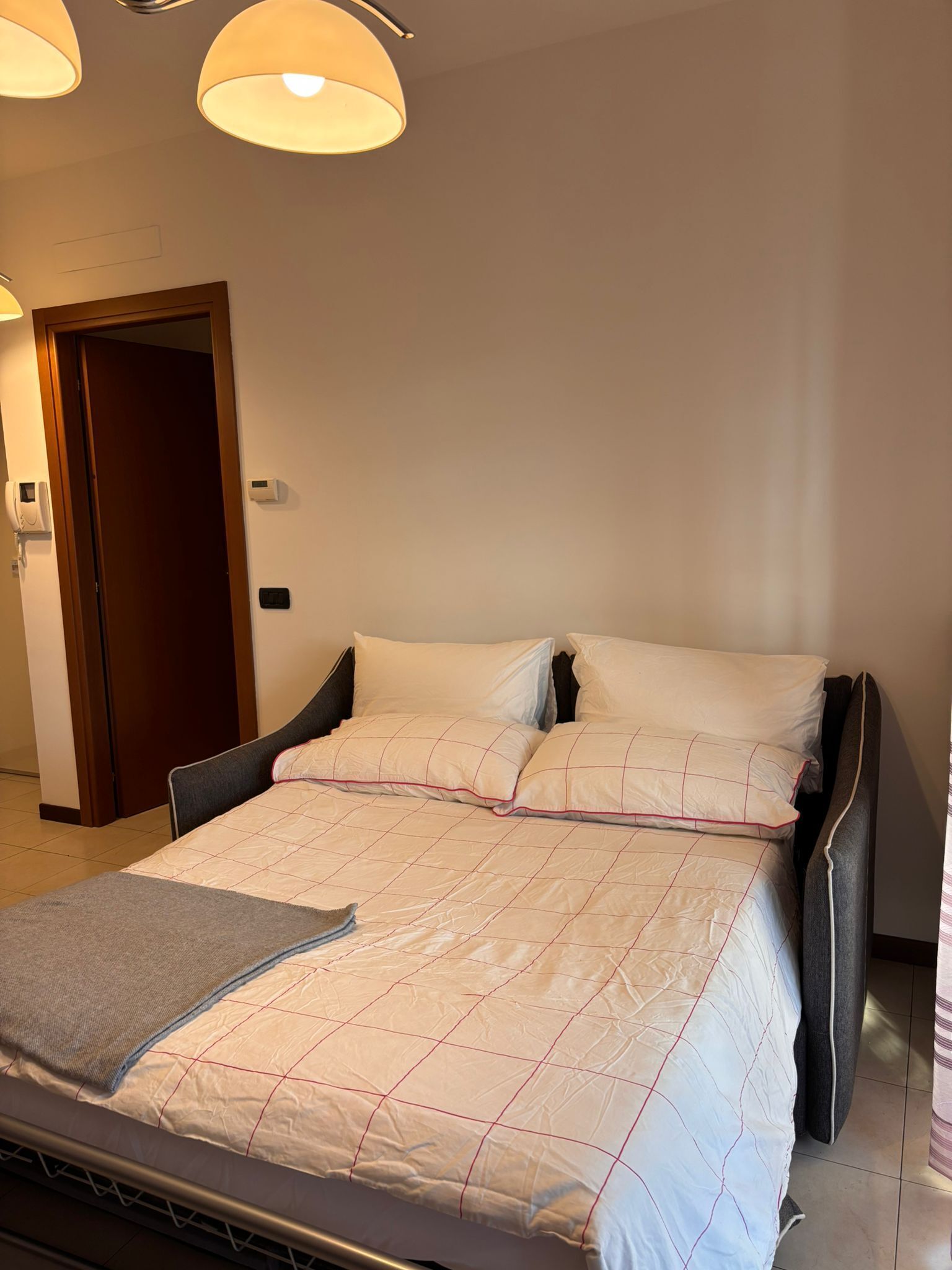 Ein ordentlich bezogenes Bett mit gesteppter Decke, ein Nachttisch mit Lampe und eine Tür zu einem anderen Raum.